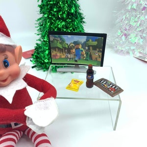 May include: A miniature scene with a small television showing a Minecraft game, a bottle of soda, a bag of crisps, and a chocolate bar on a clear table. A Christmas elf is in the foreground, with a green tinsel Christmas tree in the background.