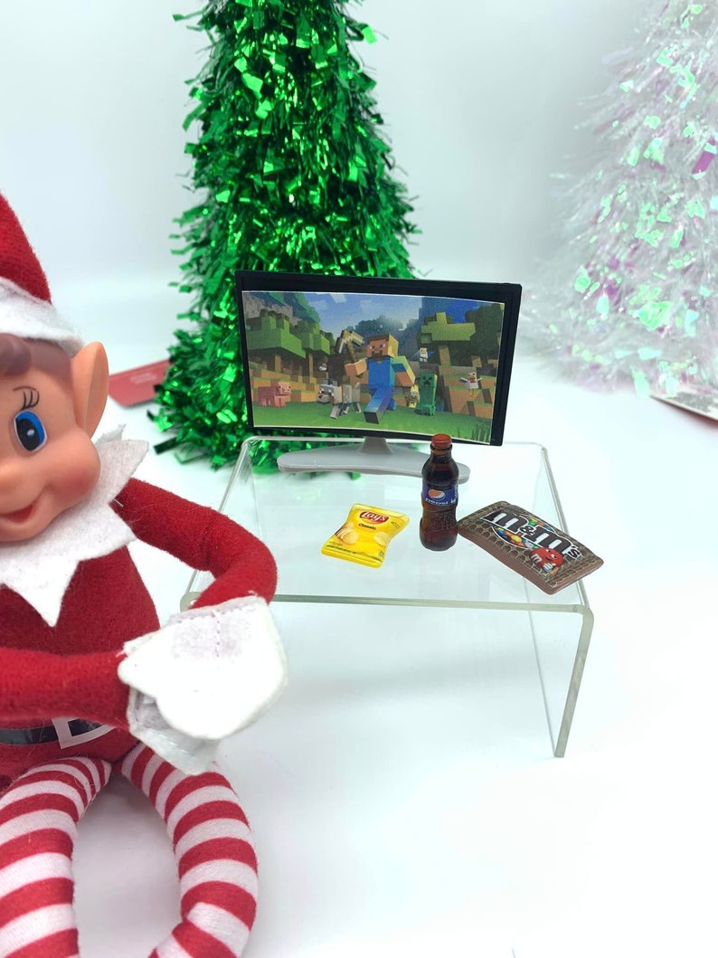 May include: A miniature scene with a small television showing a Minecraft game, a bottle of soda, a bag of crisps, and a chocolate bar on a clear table. A Christmas elf is in the foreground, with a green tinsel Christmas tree in the background.