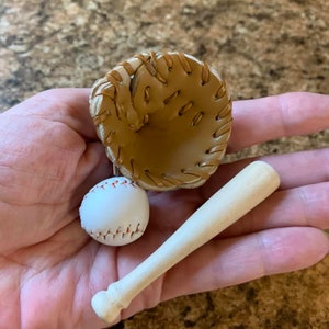 May include: A miniature baseball set consisting of a tan leather baseball glove with brown lacing, a small white baseball with red stitching, and a light-colored wooden baseball bat, all displayed together as a small collection.