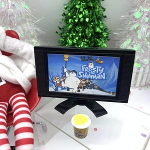 May include: A miniature black television showing the animated film "Frosty the Snowman." A small popcorn container is in front of the TV. The scene is decorated with miniature Christmas trees and a red and white striped figure.