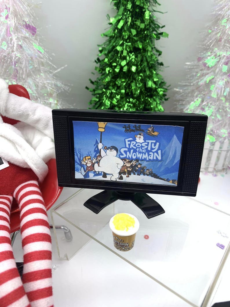 May include: A miniature black television showing the animated film "Frosty the Snowman." A small popcorn container is in front of the TV. The scene is decorated with miniature Christmas trees and a red and white striped figure.