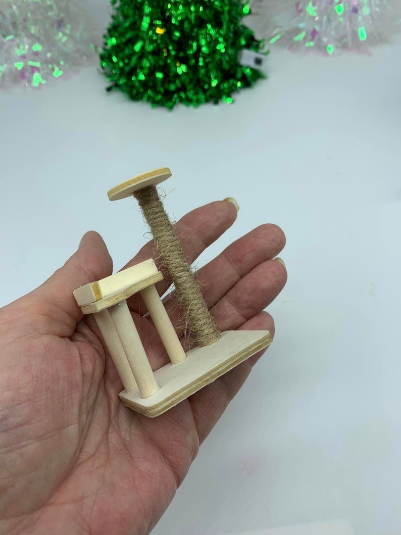 May include: A miniature wooden cat scratching post with a sisal rope scratching post and a small platform. The scratching post is held in a person's hand.