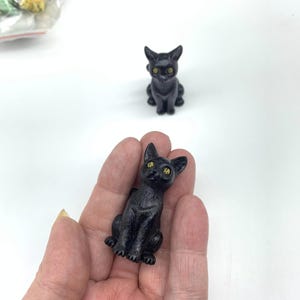 May include: Two black cat figurines with yellow eyes. The cat on the left is being held in a hand. The cat on the right is sitting on a white surface.