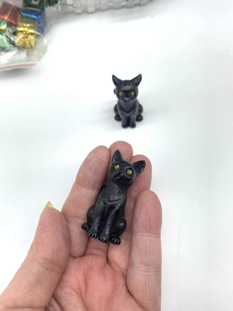 May include: Two black cat figurines with yellow eyes. The cat on the left is being held in a hand. The cat on the right is sitting on a white surface.
