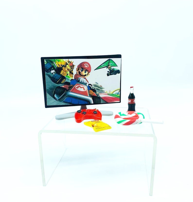 May include: A miniature gaming setup with a flat-screen monitor showing a racing game with Mario. A red controller, a yellow snack bag, a bottle of soda, and a candy cane are on a clear acrylic table. The background is white.
