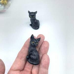 May include: A small black cat figurine with yellow eyes. The figurine is sitting on a hand.