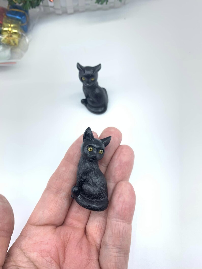 May include: A small black cat figurine with yellow eyes. The figurine is sitting on a hand.