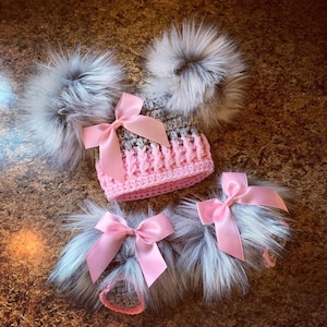May include: A handmade pink and gray crocheted hat with two large gray fur pom-poms and a pink satin bow. Two matching fur accessories with pink bows are also present. The hat and accessories are designed for babies or children.