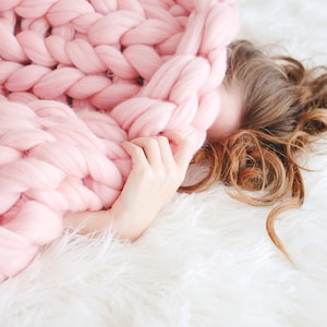 May include: A person is lying on a white fluffy blanket, partially covered by a large, pink knitted blanket. The blanket is made of thick, chunky yarn and has a soft, cosy texture.