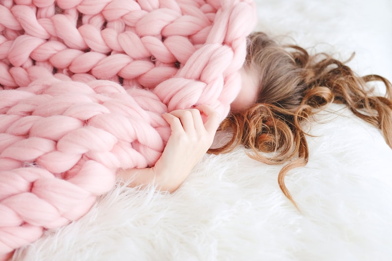 May include: A person is lying on a white fluffy blanket, partially covered by a large, pink knitted blanket. The blanket is made of thick, chunky yarn and has a soft, cosy texture.