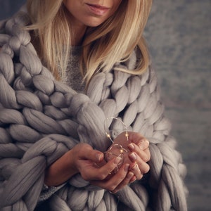 May include: A person wrapped in a chunky knit grey blanket holds a strand of fairy lights in their hands.