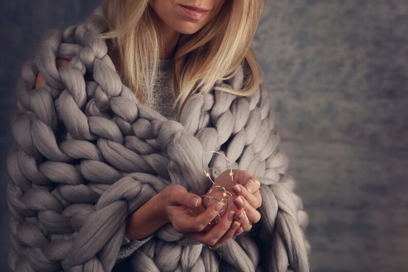 May include: A person wrapped in a chunky knit grey blanket holds a strand of fairy lights in their hands.