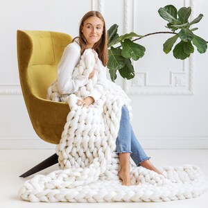 May include: A woman sits in a yellow armchair, wrapped in a large, white chunky knit blanket. The blanket is draped over her legs and extends out onto the floor.