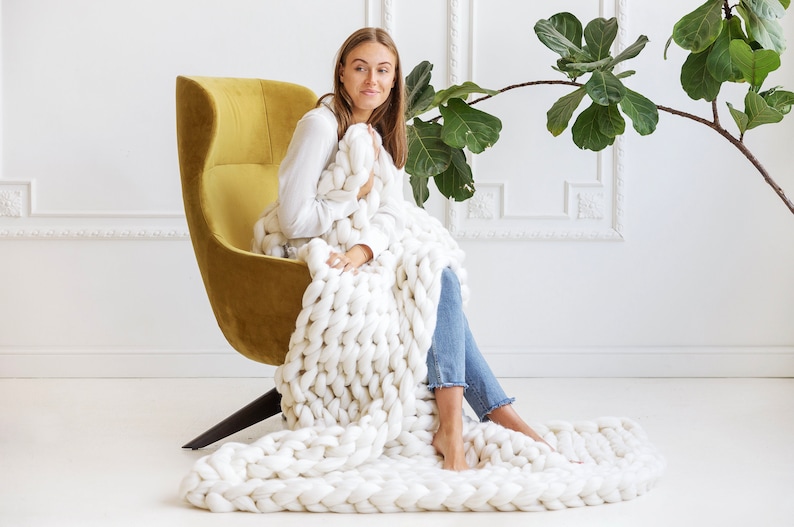 May include: A woman sits in a yellow armchair, wrapped in a large, white chunky knit blanket. The blanket is draped over her legs and extends out onto the floor.