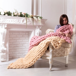 May include: A woman sits in a white armchair, wrapped in a chunky knit throw with pink and cream stripes. The throw is draped over the chair and the woman's legs. The room is decorated with white walls and a white fireplace.