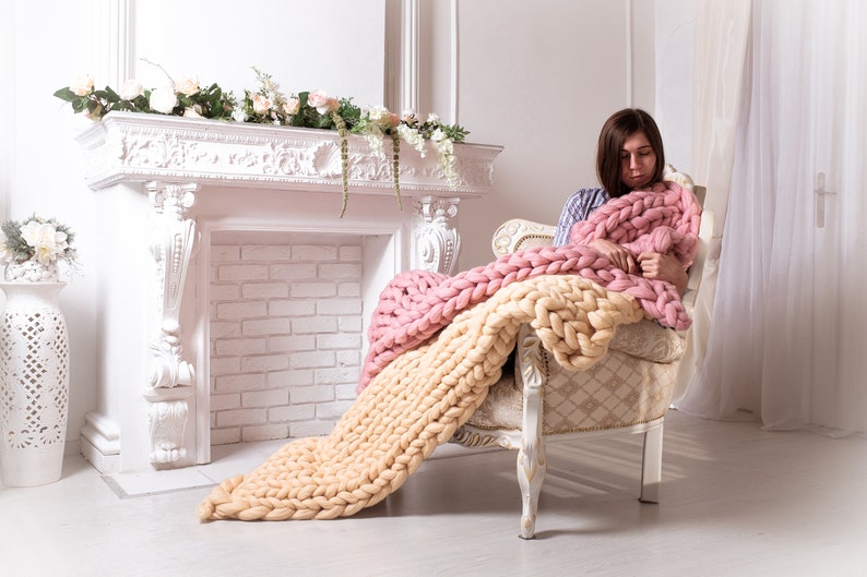 May include: A woman sits in a white armchair, wrapped in a chunky knit throw with pink and cream stripes. The throw is draped over the chair and the woman's legs. The room is decorated with white walls and a white fireplace.