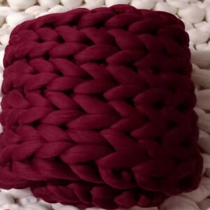 May include: A chunky knit throw blanket in a deep burgundy colour. The blanket is made of thick yarn and has a soft, cosy texture.