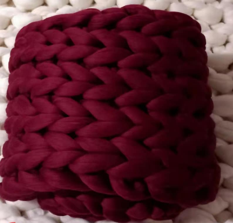 May include: A chunky knit throw blanket in a deep burgundy colour. The blanket is made of thick yarn and has a soft, cosy texture.