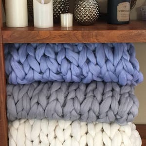 May include: Three chunky knit blankets stacked on a wooden shelf. The top blanket is light blue, the middle blanket is grey, and the bottom blanket is white.