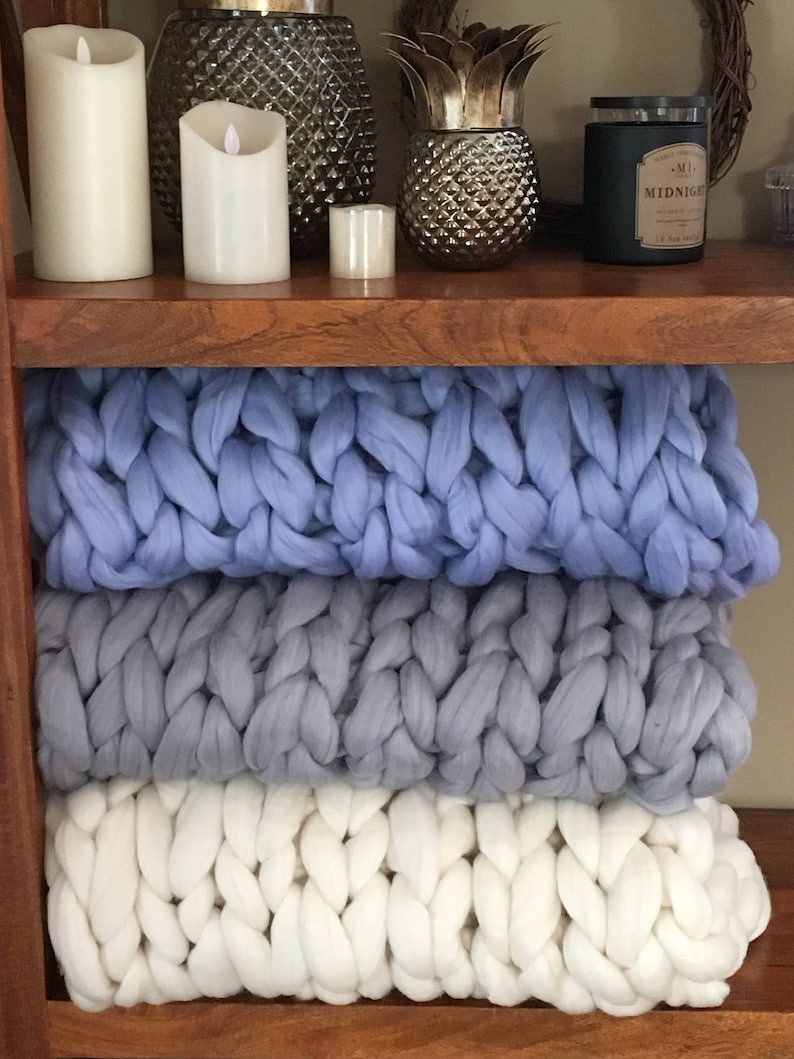 May include: Three chunky knit blankets stacked on a wooden shelf. The top blanket is light blue, the middle blanket is grey, and the bottom blanket is white.