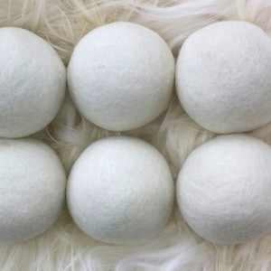 May include: Six white wool dryer balls, arranged in a circle on a white fluffy surface.