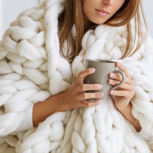 May include: A person wrapped in a large, white, chunky knit blanket holds a grey ceramic mug.