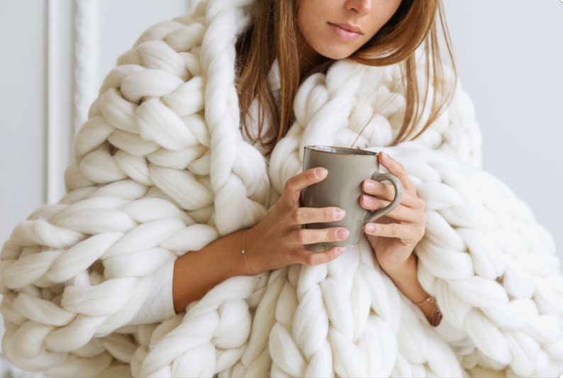 May include: A person wrapped in a large, white, chunky knit blanket holds a grey ceramic mug.