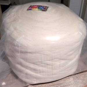 May include: A large, cylindrical bundle of white fiber, tightly wrapped in clear plastic. A small, colorful label is visible on top. The fiber appears soft and fluffy, suggesting a textile or craft material.