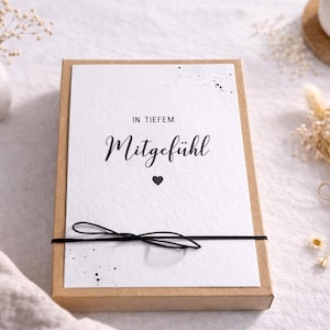 Könnte beinhalten: Eine rechteckige Geschenkbox mit einer weißen Karte mit den Worten "IN TIEFEM Mitgefühl" und einem Herzsymbol. Die Box ist mit einem schwarzen Band gebunden. Trockenblumen und eine Kerze im Hintergrund.