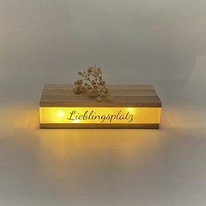 Könnte beinhalten: Eine Holzkiste mit einem Licht im Inneren, das den Text "Lieblingsplatz" beleuchtet. Die Kiste steht auf einer weißen Oberfläche mit einem kleinen Strauß getrockneter Blumen darauf.