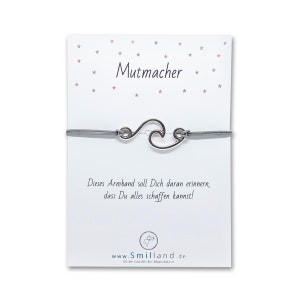 Könnte beinhalten: Ein graues Armband mit einem silbernen Wellen-Anhänger und dem Text "Mutmacher" auf einer weißen Karte mit rosa und grauen Sternen. Die Karte enthält auch den Text "Dieses Armband soll Dich daran erinnern, dass Du alles schaffen kannst!" und "www.Smilland.de Deine nordische Manufaktur."