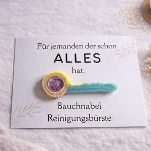 Könnte beinhalten: Eine Grußkarte mit deutschem Text und einer handgefertigten Bauchnabelreinigungsbürste. Die Bürste ist gelb, lila und blau. Die Karte liegt auf einer weißen Oberfläche mit kleinen Blumengestecken.