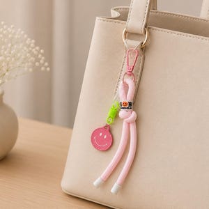 Könnte beinhalten: Eine beige Handtasche mit einem dekorativen Schlüsselanhänger. Das Accessoire besteht aus einem geknoteten rosa Seil, einem lindgrünen Spiralelement und einem runden rosa Anhänger mit weißem Smiley-Gesicht. Der Text "smile" ist in Schwarz.