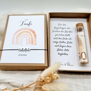 Pode incluir: Uma caixa castanha com um cart&atilde;o branco no seu interior. O cart&atilde;o tem um desenho de arco-&iacute;ris e o texto "208 Taufe Leni 22.05.2024". A caixa tamb&eacute;m cont&eacute;m um tubo de vidro com uma nota de 50 euros no seu interior. O texto no cart&atilde;o diz: "Como o arco-&iacute;ris &eacute; o sinal da alian&ccedil;a entre Deus e a terra, que o amor e a b&ecirc;n&ccedil;&atilde;o de Deus acompanhem sempre a sua vida."