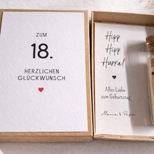 以下が含まれることがあります： 「ZUM 18. HERZLICHEN GL&Uuml;CKWUNSCH」と赤いハートのカードが入ったギフトボックス。内側には「Hipp Hipp Hurra！」と紙幣が入ったガラス管が入ったカードが入っています。