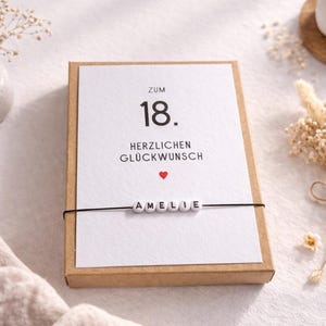 以下が含まれることがあります： 「Zum 18. Herzlichen Gl&uuml;ckwunsch」の文字と赤いハートが描かれたバースデーカード。名前「AMELIE」は、白い文字ビーズで黒い紐に綴られています。カードは茶色の箱に入っており、背景にはドライフラワーとキャンドルがあります。