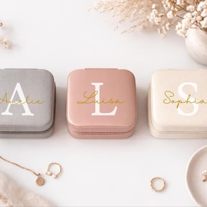 Könnte beinhalten: Drei quadratische Schmuckkästchen in Grau, Rosa und Creme. Jede Box hat einen großen Anfangsbuchstaben und einen Namen in goldener Schrift. Die Boxen stehen auf einer weißen Oberfläche mit Schmuck und dekorativen Blumen.