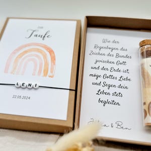 Pode incluir: Uma caixa marrom com um cart&atilde;o branco dentro. O cart&atilde;o tem um arco-&iacute;ris de aquarela com a palavra "Taufe" e o nome "LENI" escrito com contas. A data "22.05.2024" est&aacute; impressa abaixo das contas. A caixa tamb&eacute;m cont&eacute;m um pequeno tubo de vidro com uma nota enrolada dentro.
