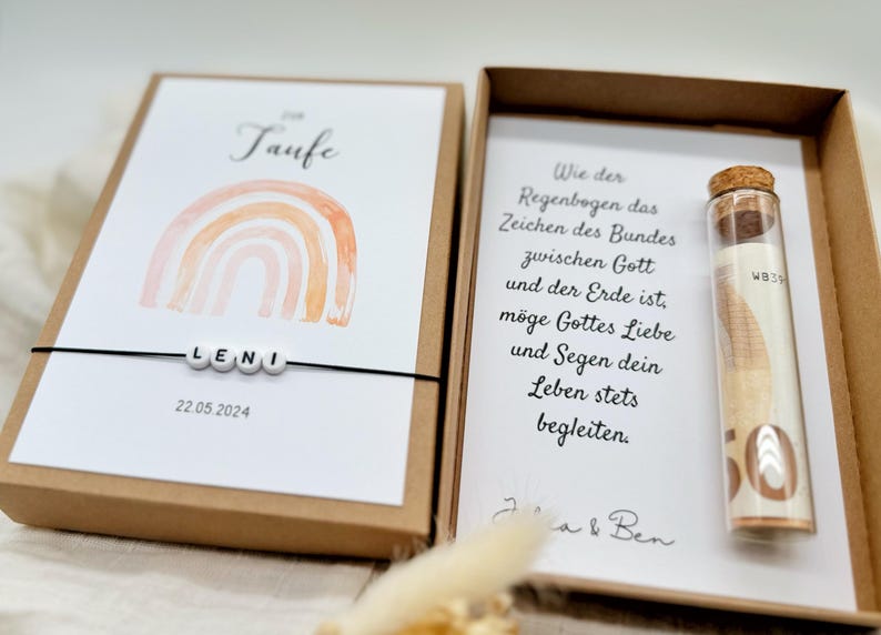 Pode incluir: Uma caixa marrom com um cart&atilde;o branco dentro. O cart&atilde;o tem um arco-&iacute;ris de aquarela com a palavra "Taufe" e o nome "LENI" escrito com contas. A data "22.05.2024" est&aacute; impressa abaixo das contas. A caixa tamb&eacute;m cont&eacute;m um pequeno tubo de vidro com uma nota enrolada dentro.