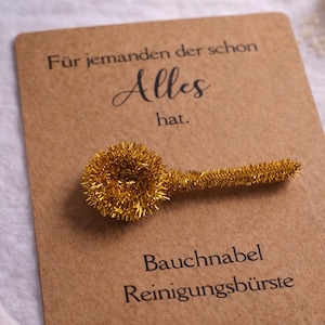 Könnte beinhalten: Eine goldfarbene Bauchnabelreinigungsbürste liegt auf einer braunen Karte mit deutschem Text. Die Karte trägt auch den Text "Alles hat." und "Bauchnabel Reinigungsbürste." Die Bürste besteht aus einem flauschigen Material. Kleine weiße Blumen sind im Hintergrund.