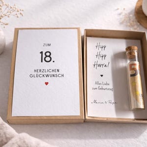 以下が含まれることがあります： 「Zum 18. Herzlichen Gl&uuml;ckwunsch」と書かれたカードと、数字の5が入った小さなガラス瓶が入ったギフトボックス。カードには赤いハートも描かれています。ボックスは白い表面に置かれています。