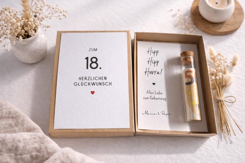 以下が含まれることがあります： 「Zum 18. Herzlichen Gl&uuml;ckwunsch」と書かれたカードと、数字の5が入った小さなガラス瓶が入ったギフトボックス。カードには赤いハートも描かれています。ボックスは白い表面に置かれています。