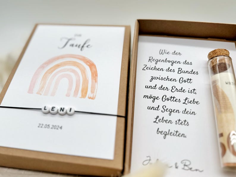 Pode incluir: Um cart&atilde;o branco com um arco-&iacute;ris de aquarela e o texto "Taufe" e "LENI" com a data "22.05.2024". O cart&atilde;o est&aacute; em uma caixa marrom com um cart&atilde;o branco dentro que tem texto sobre um arco-&iacute;ris como sinal do amor e da b&ecirc;n&ccedil;&atilde;o de Deus. Um pequeno frasco de vidro com um peda&ccedil;o de papel enrolado dentro tamb&eacute;m est&aacute; na caixa.