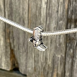 Może przedstawiać: Srebrny charms w kształcie buta kowbojskiego. But ma szczegółowe szwy i klamrę. Charms jest nawleczony na srebrny łańcuszek, z drewnianym tłem.