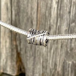 May include: A silver charm bead with a feather design. The bead is cylindrical with a series of curved lines and a feather detail. It is strung on a silver snake chain, perfect for bracelets or necklaces.