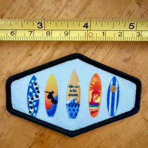 Op de afbeelding: Een zeshoekige geborduurde patch met vijf surfplanken op een lichtblauwe achtergrond, geplaatst onder een geel meetlint dat een breedte van ongeveer 9 cm aangeeft. De surfplanken tonen diverse patronen, waaronder een silhouet van een surfer en de tekst "take me to the ocean" op de middelste plank.