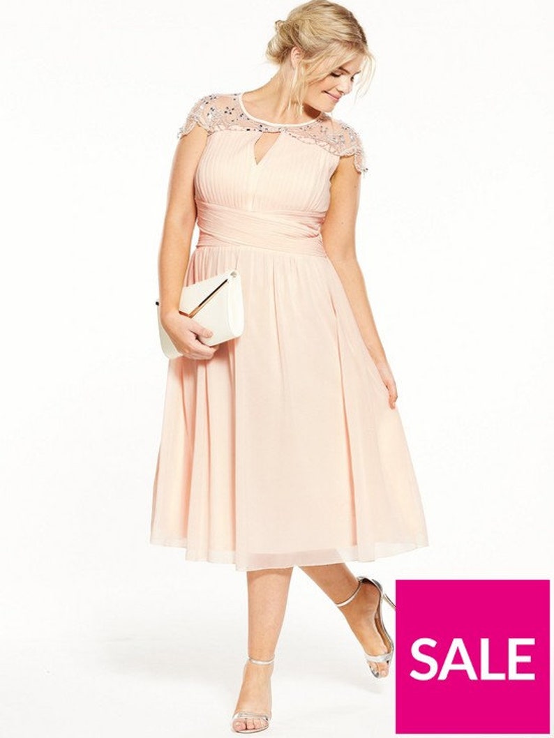 May include: A blush-colored knee-length dress with a pleated bodice and a keyhole neckline. The dress features cap sleeves with beaded embellishments and a wrapped waist. A white clutch and silver heels complete the look. The image includes a pink "SALE" graphic.