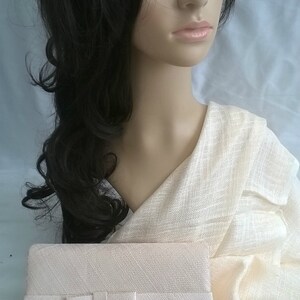 May include: A light beige clutch with a bow detail, resting on a matching fabric. The clutch has a rectangular shape. The background features a mannequin with dark hair and a cream-colored shawl.