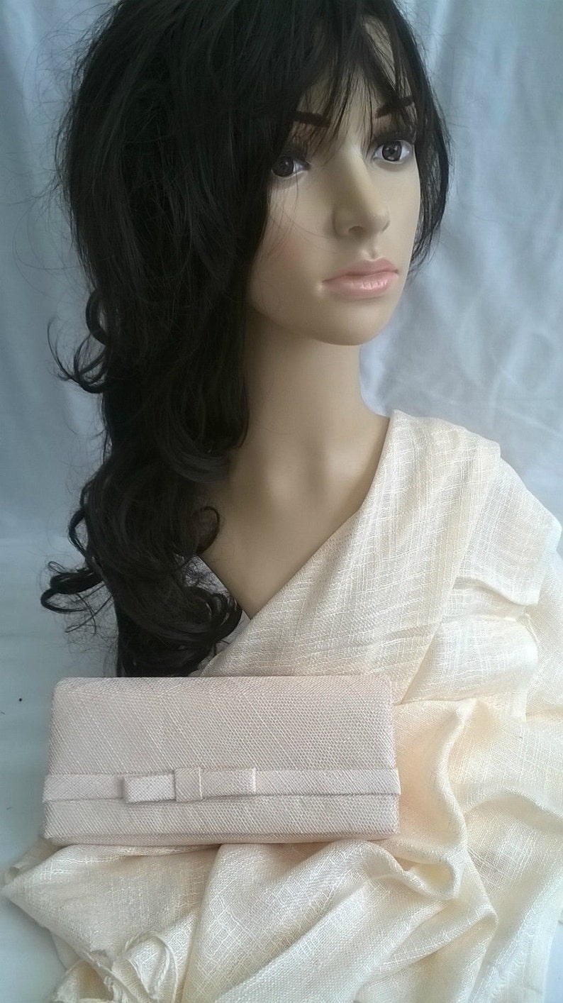 May include: A light beige clutch with a bow detail, resting on a matching fabric. The clutch has a rectangular shape. The background features a mannequin with dark hair and a cream-colored shawl.