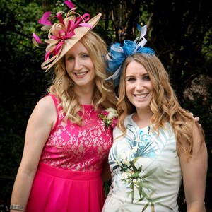 May include: Two women wearing formal attire. One wears a pink dress with a lace bodice and a beige hat with pink and beige accents. The other wears a white dress with a floral print and a blue headpiece.